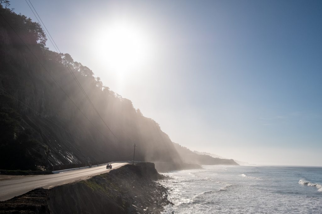 De zon schijnt over een bergkam die afloopt naar de kust. In de verte rijdt een auto over een weg.